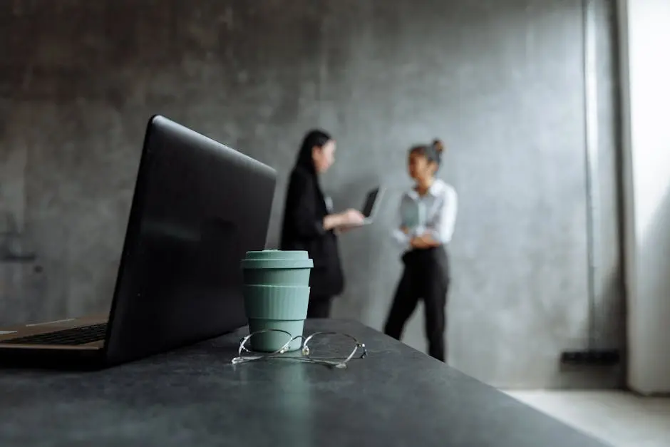 Two professionals discuss business strategies in a stylish office setting with a focus on a laptop and glasses.