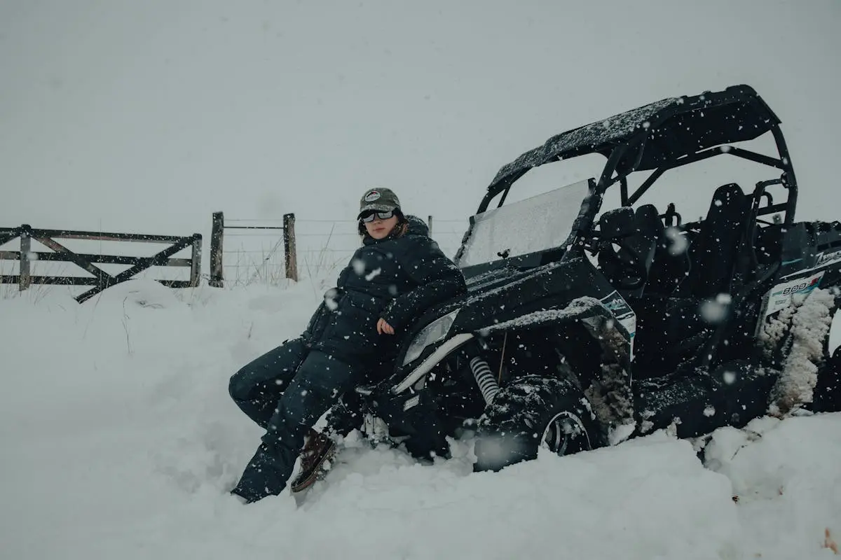 A person wearing winter clothing leans on a utility vehicle during a heavy snowfall. Outdoor winter scene.