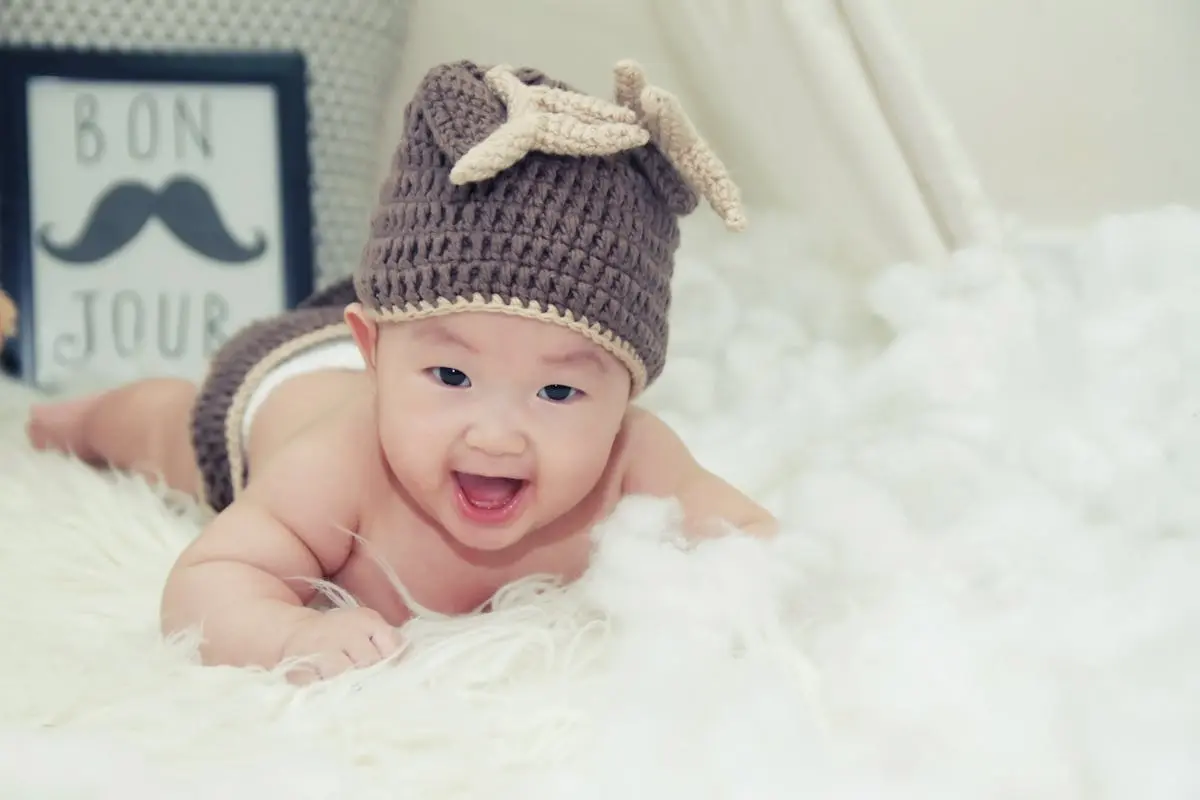 A joyful baby wearing a brown knitted hat lies on a soft surface, radiating happiness.