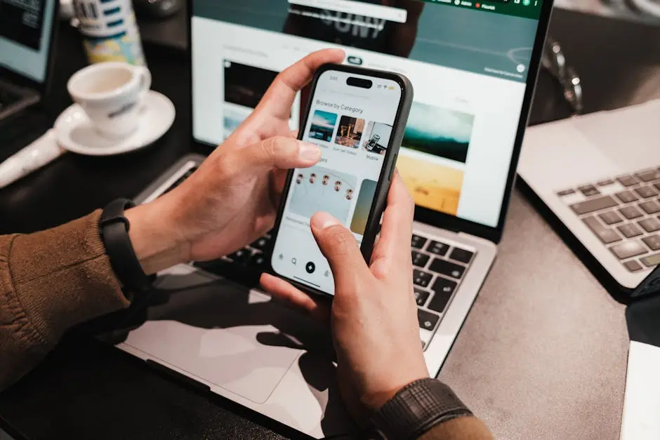 Close-up of hands using a smartphone over a laptop in a modern office environment.