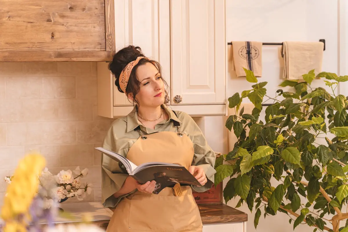 Woman with bandana and apron reads a book in a warmly lit, plant-filled kitchen.woman with plants eco friendly cleaning green products natural toxic free no harsh chemicals