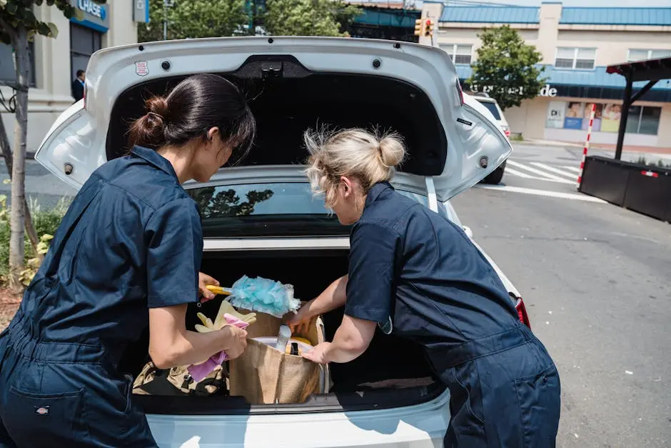 Two women in uniforms unloading cleaning supplies from a car trunk in an urban area.