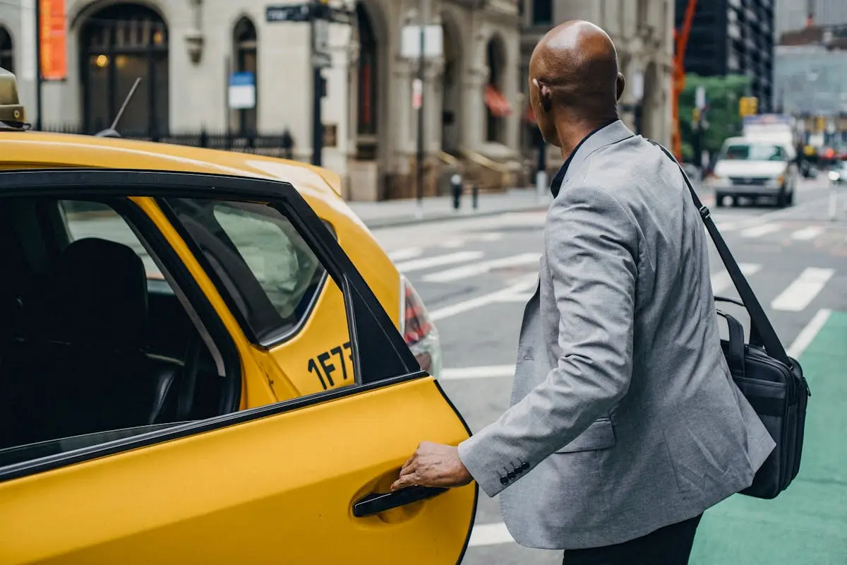 A businessman in a gray suit opens the door of a yellow taxi on a bustling city street, ready for his daily commute wearing a utility backpack with his computer