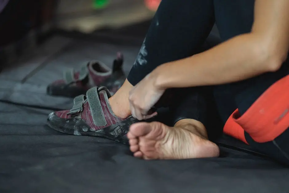 Side view of unrecognizable barefoot mountaineer sitting on mat with crossed legs while putting on rock climbing shoes during training in gym on blurred background