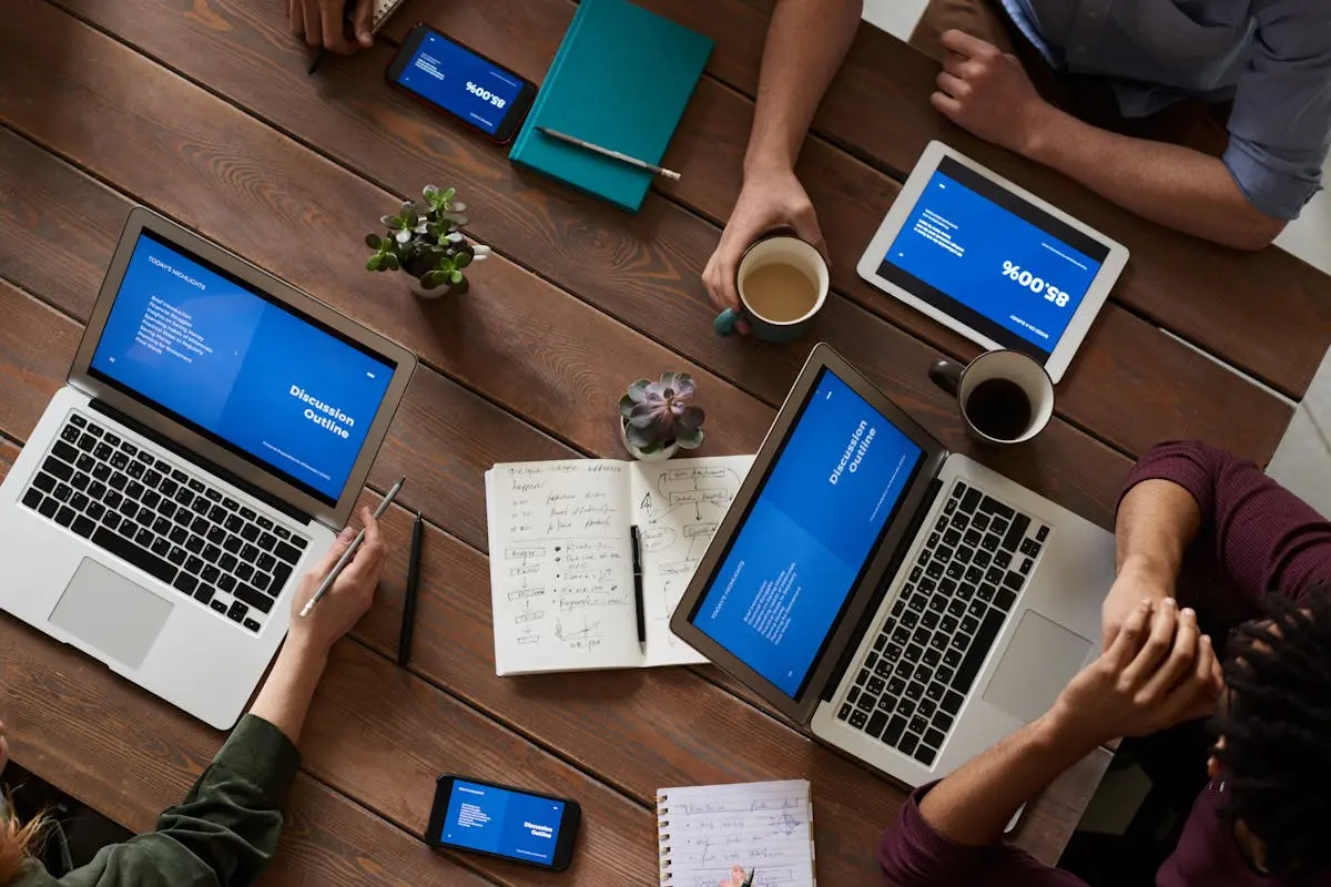 Top view of a diverse team using technology in a meeting. Laptops, tablets, and smartphones on a wooden table.