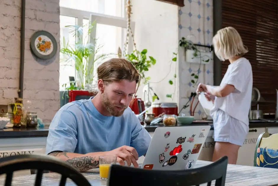 Man working on a laptop in a communal kitchen while woman prepares food in the background.