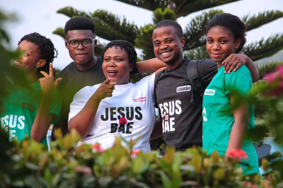 A cheerful group of friends posing in ‘Jesus is Bae’ shirts amidst greenery.