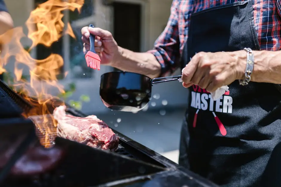 A person grilling steak outdoors with flames, wearing an apron, adding sauce.