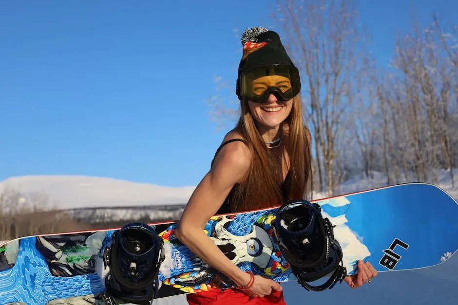 Smiling woman in beanie and goggles holding snowboard in a sunny winter scene.