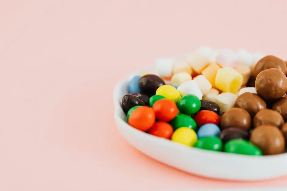 A colorful assortment of candies in a white bowl, featuring chocolates and gummies on a pastel pink background.