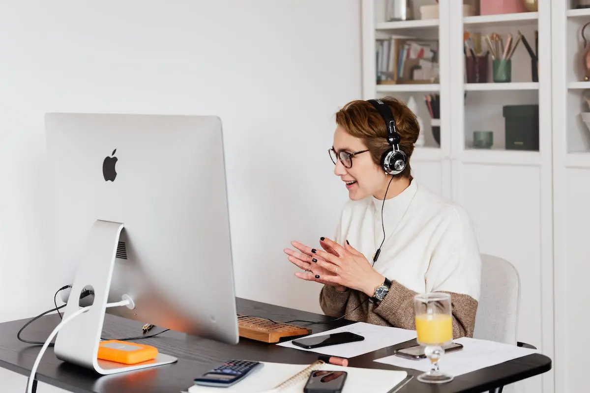 Cheerful female in casual wear and wired headphones having conversation via video call on computer while working in light modern workplace