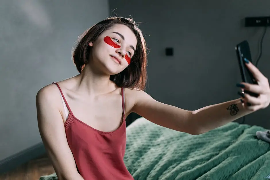 A young woman applying under-eye patches while taking a selfie indoors, illustrating a self-care routine.