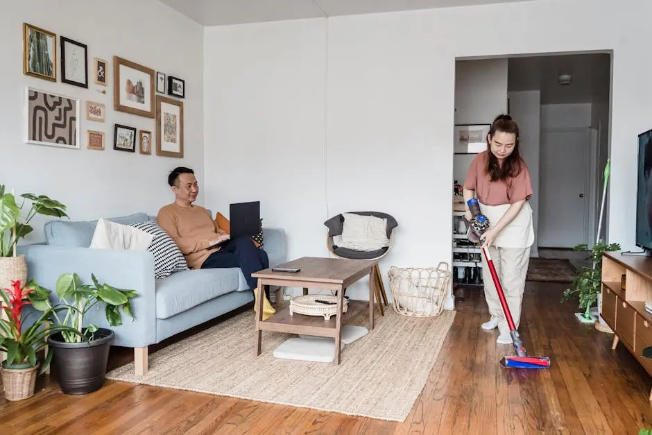 A man working on a laptop while a woman vacuums the living room, showcasing modern lifestyle balance.