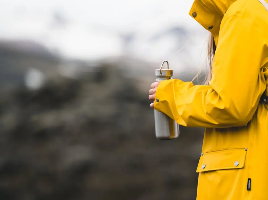 A person in a yellow raincoat holds a metal water bottle, set against a blurred Icelandic landscape.