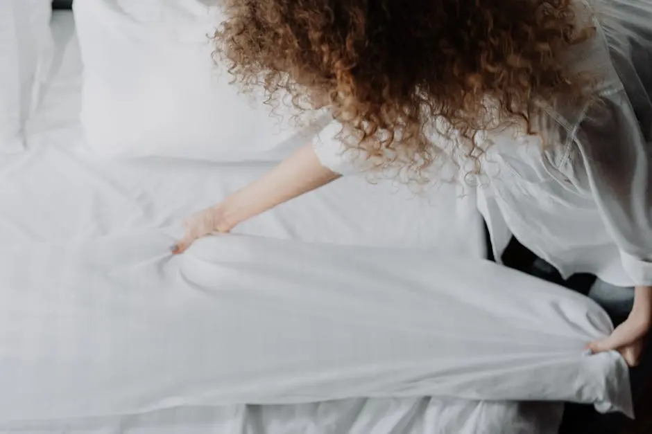 A person arranging white bed linens in a serene bedroom, focusing on clean and cozy atmosphere.