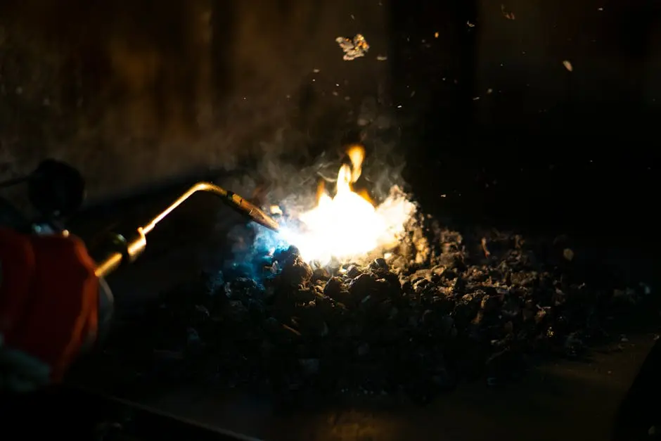 Close-up of a torch flame heating charcoal, emitting smoke in an industrial setting.