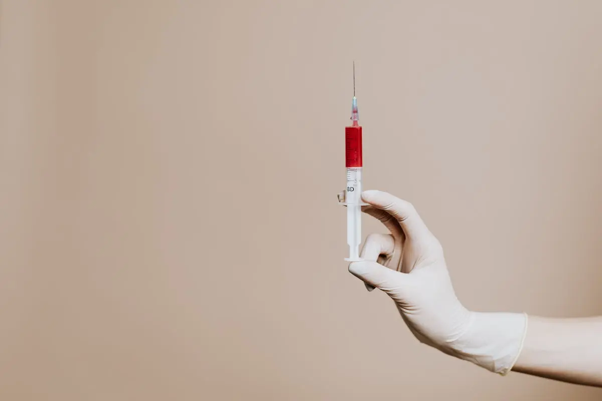 Close-up of a gloved hand holding a syringe with red liquid against neutral background.