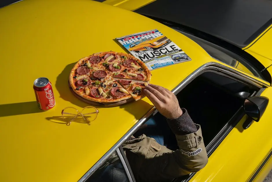 Pizza, cola, and a magazine atop a vintage yellow car hood create a casual lifestyle scene.