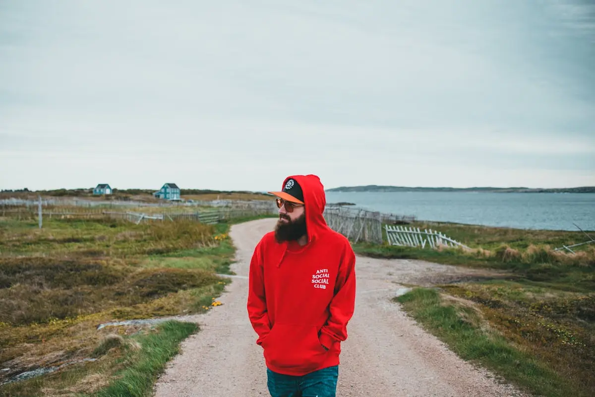 Bearded Man in Red Hoodie Standing on Dirt Road between Grasses