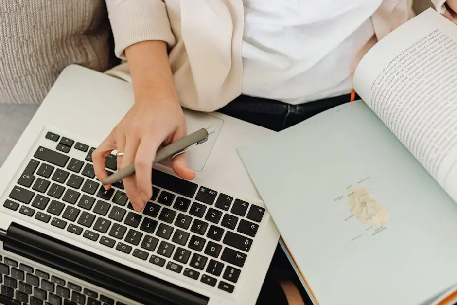 Close-up of a person working on a laptop and reading an open book. Ideal for educational themes.