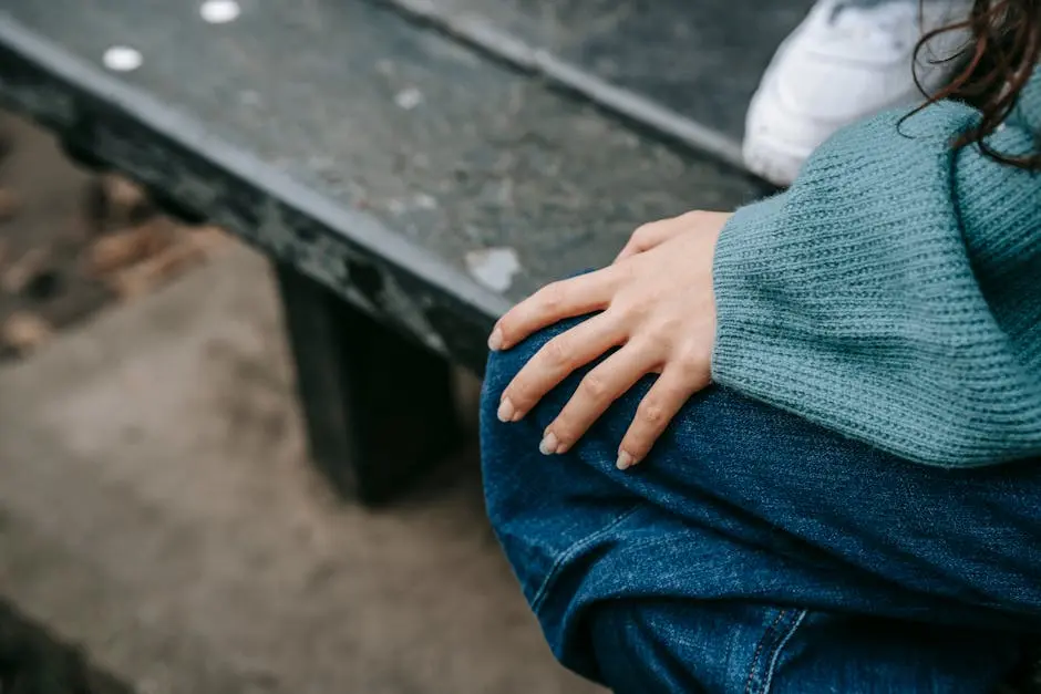 From above of anonymous female in casual outfit sitting on bench with hand on knee while resting on street with unrecognizable person on blurred background