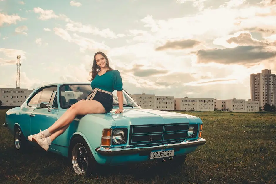 Young woman sitting on a classic blue car in a grassy urban area, enjoying a sunny day.