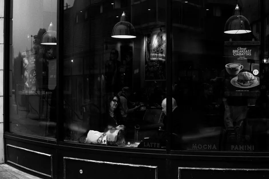 Grayscale image of a bustling urban café with people chatting and enjoying drinks through a glass wall.