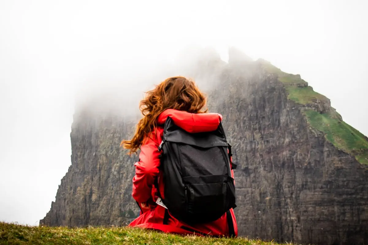 A woman sits with a Rucksack backpacks in front of fog-covered cliffs in the Faroe Islands, perfect for adventure travel themes.