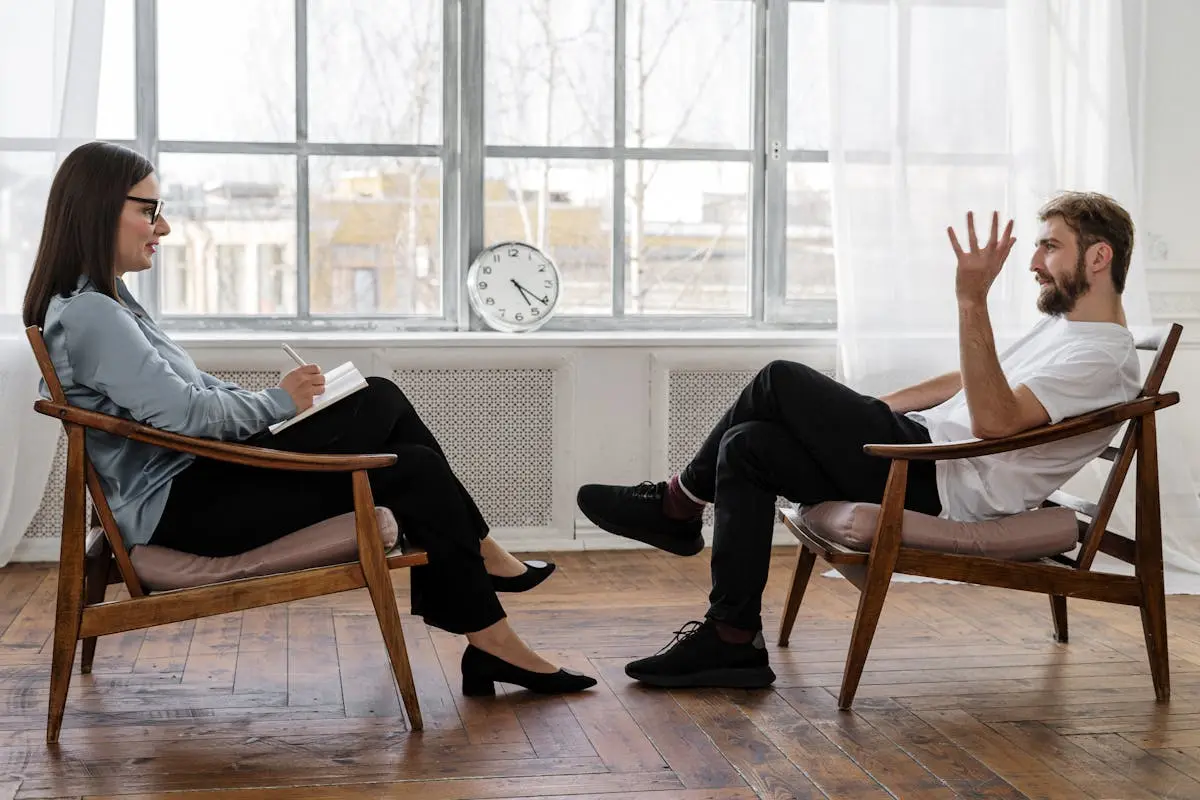 Person in Black Pants and Black Shoes Sitting on Brown Wooden Chair