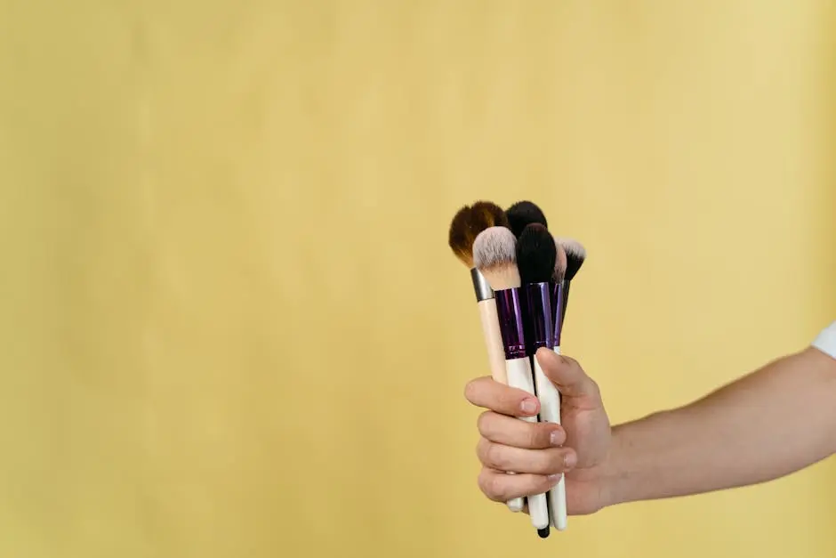 Close-Up Shot of a Person Holding Makeup Brushes on Pale Yellow Background