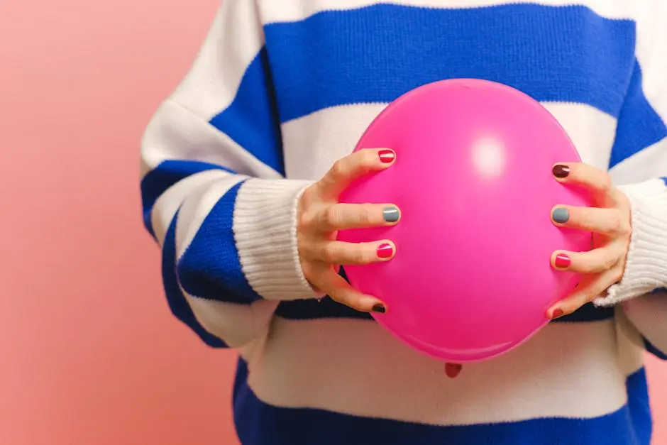 Close-up of hands with colorful nails holding a pink balloon, wearing a blue and white striped sweater against a pink background.