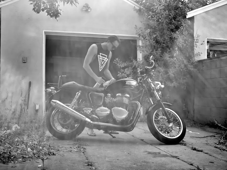 A man cleans a motorcycle in a sunlit driveway. Black and white photography enhances the classic look.