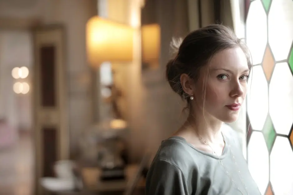 Pretty casual serious lady with hairstyle intently looking at camera while standing near stained glass window in bright luxury room of old mansion