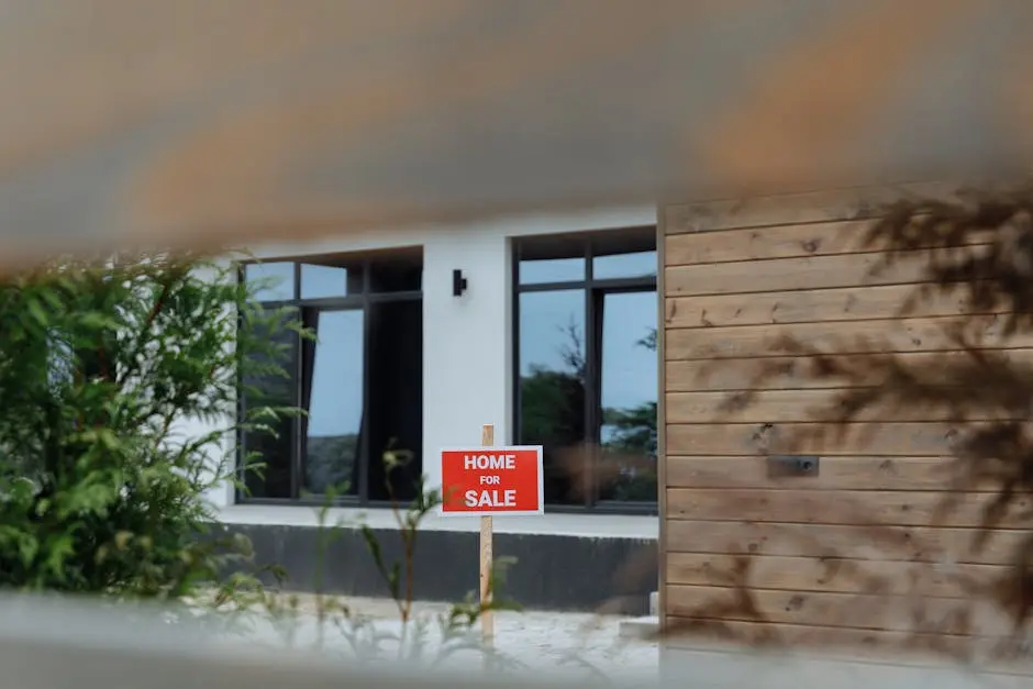 A contemporary home with a ‘For Sale’ sign in the front yard, framed by greenery.