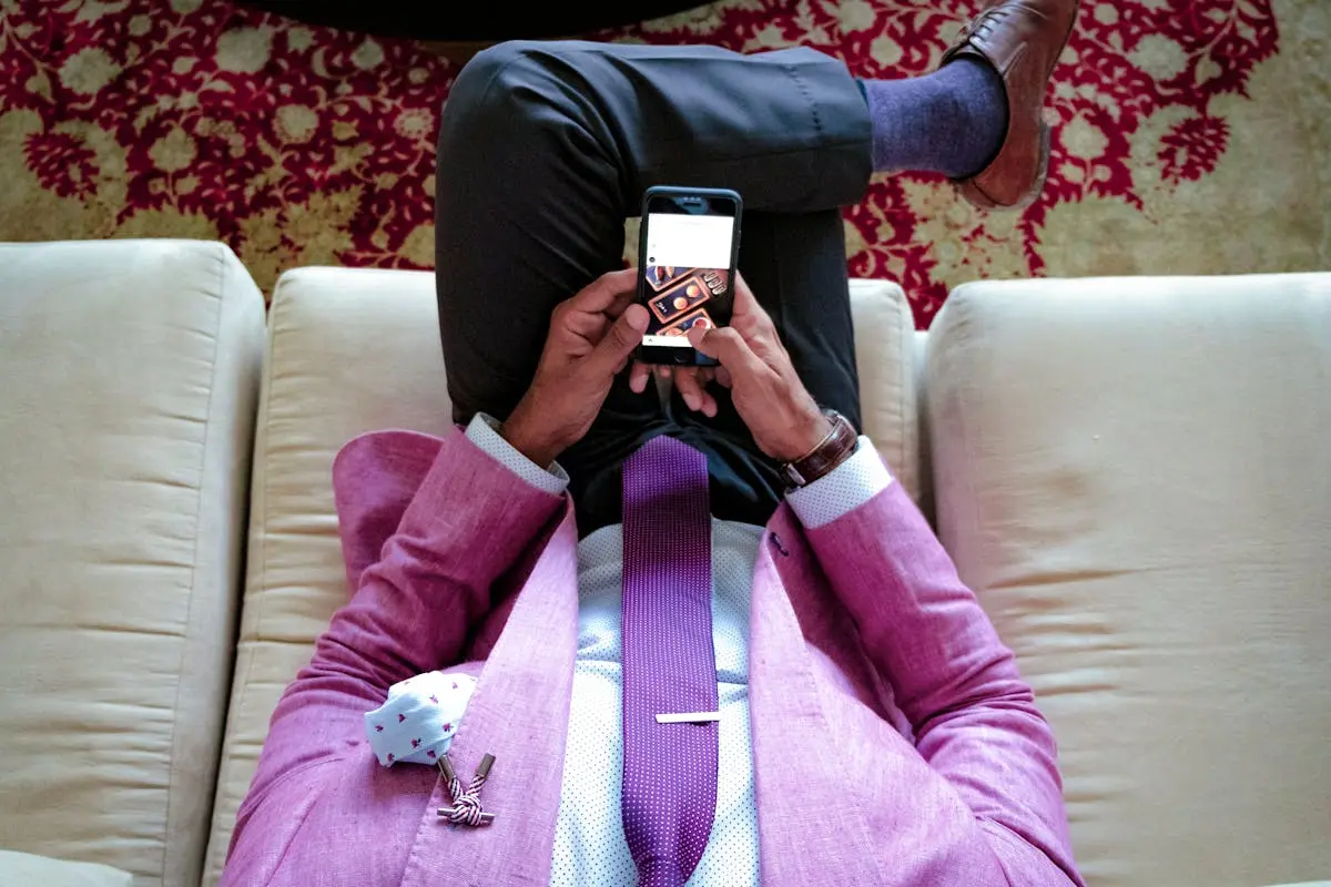 A man in a stylish pink suit sits on a sofa using a smartphone, showcasing modern business fashion.