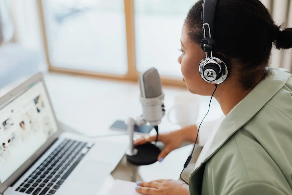 Black woman wearing headphones recording podcast with laptop and microphone indoors.