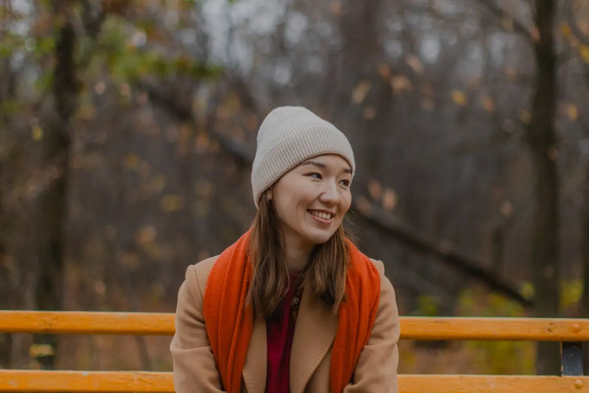 Smiling woman in warm clothing seated on a bench amidst fall foliage.