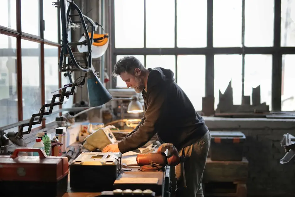 A dedicated craftsman working intently by a large window in a sunlit workshop filled with tools.