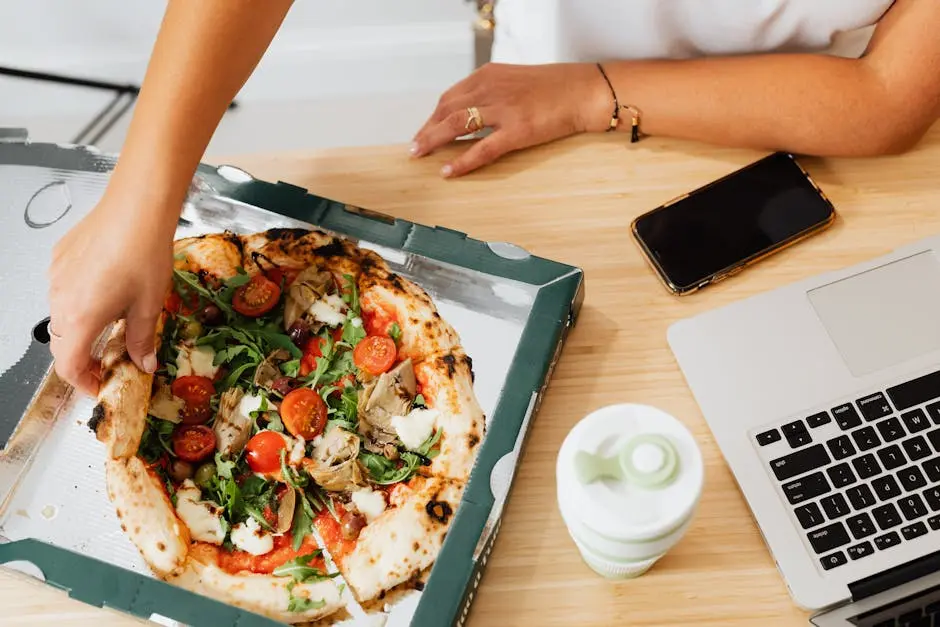 Hand reaching for a slice of pizza during an office lunch break with laptop and smartphone nearby.