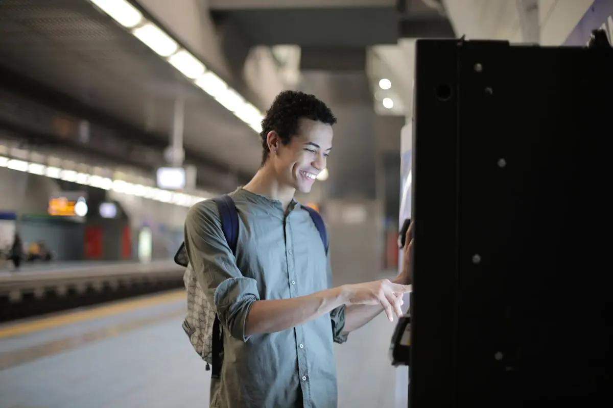 Joyful young African American male in casual clothes with backpack focusing and interacting with vending machine at underground station against blurred railway platform