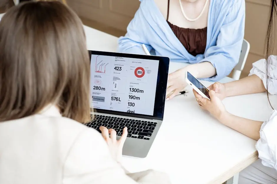 A group of women collaborating at a desk using a laptop and smartphone for business tasks indoors.