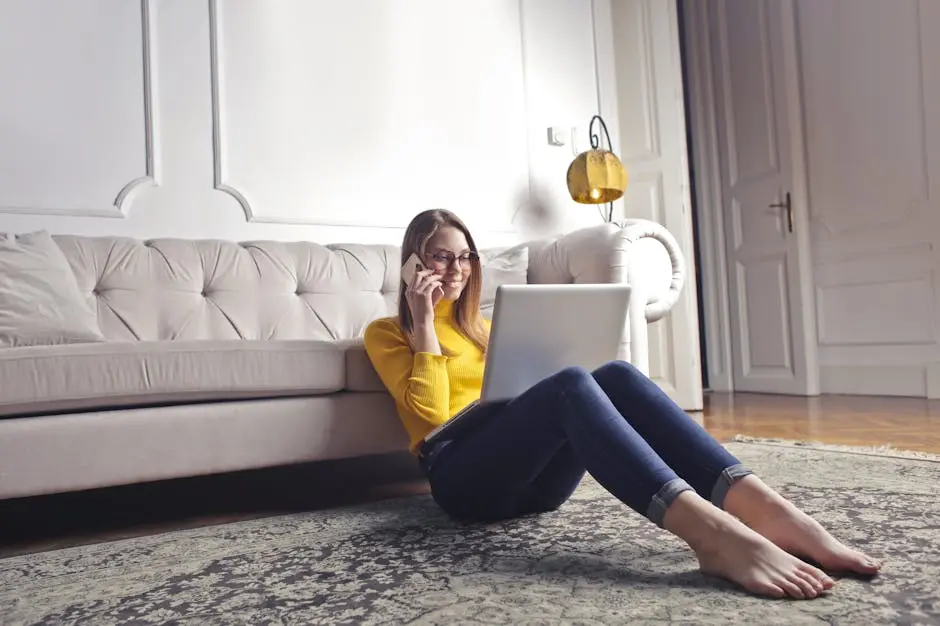 Femme souriante, pieds nus, portant des lunettes et des vêtements décontractés, utilisant un ordinateur portable et passant un appel téléphonique, assise sur le sol, appuyée sur le canapé et travaillant sur son ordinateur portable, dans un intérieur luxueux de salon clair