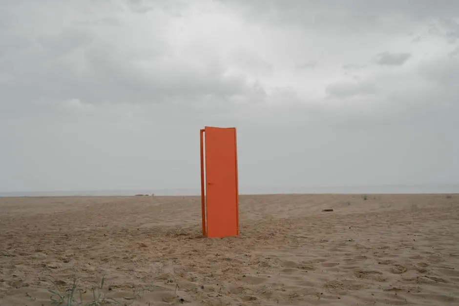 An orange door stands alone on an empty beach under a cloudy sky.