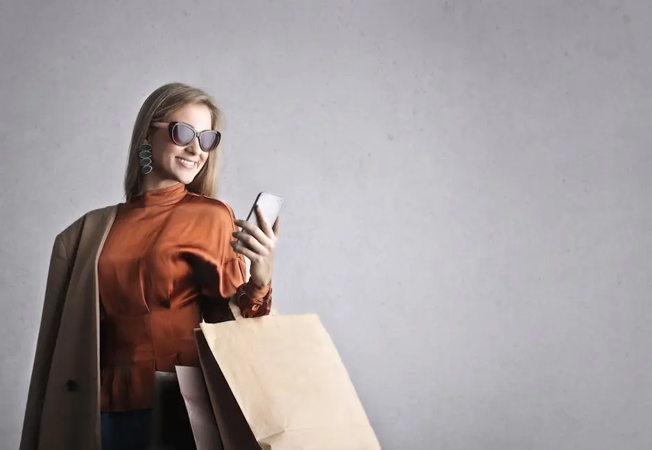 Smiling female in trendy wear and stylish sunglasses standing with paper shopping bags on background of gray wall and browsing mobile phone