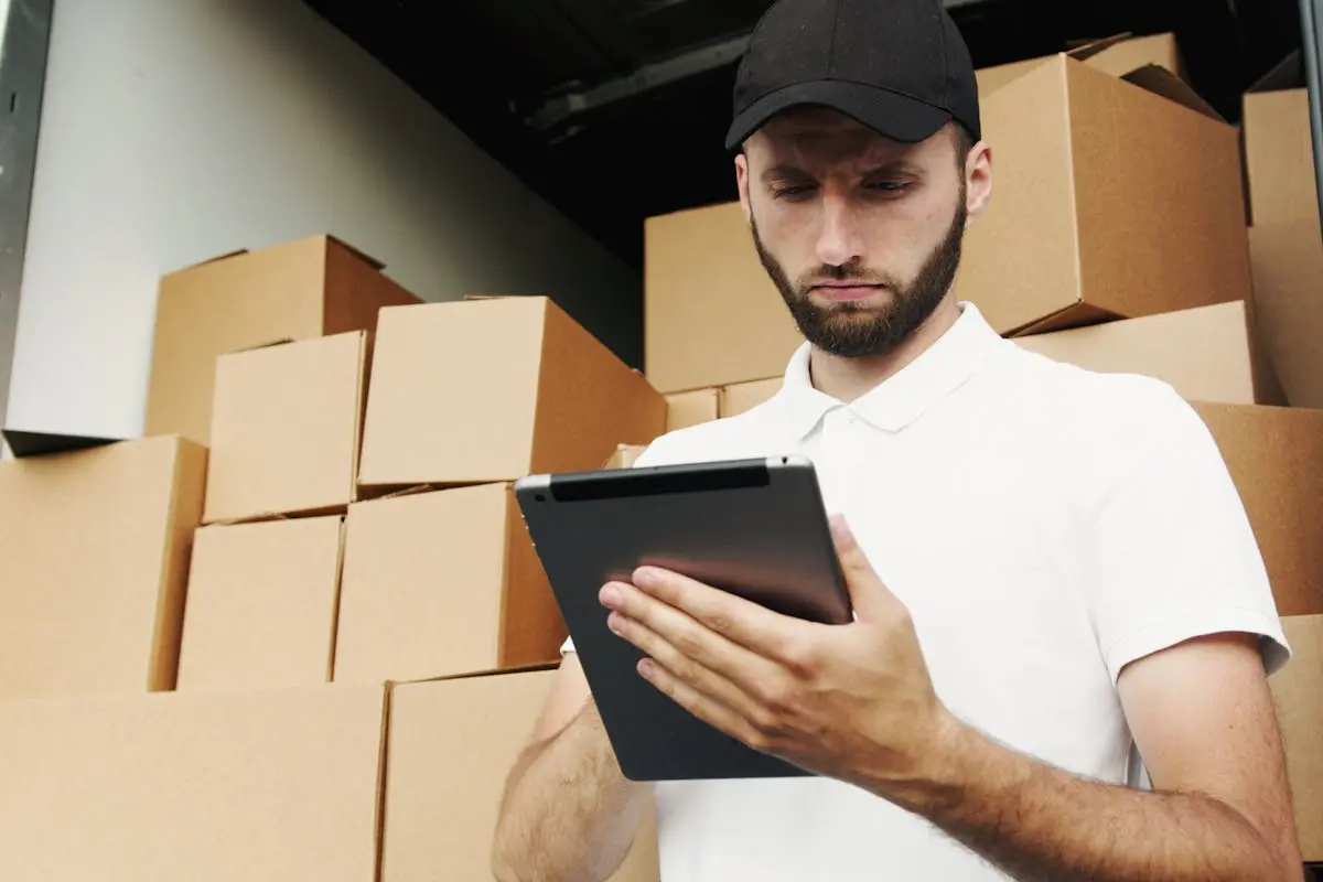 Delivery worker using a tablet to manage shipments with stacked boxes in the background.