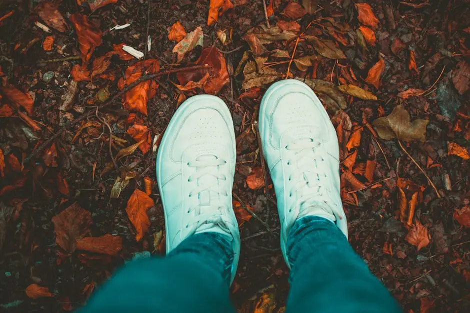 Person wearing white sneakers standing on a path covered with autumn leaves, showcasing a fall vibe.