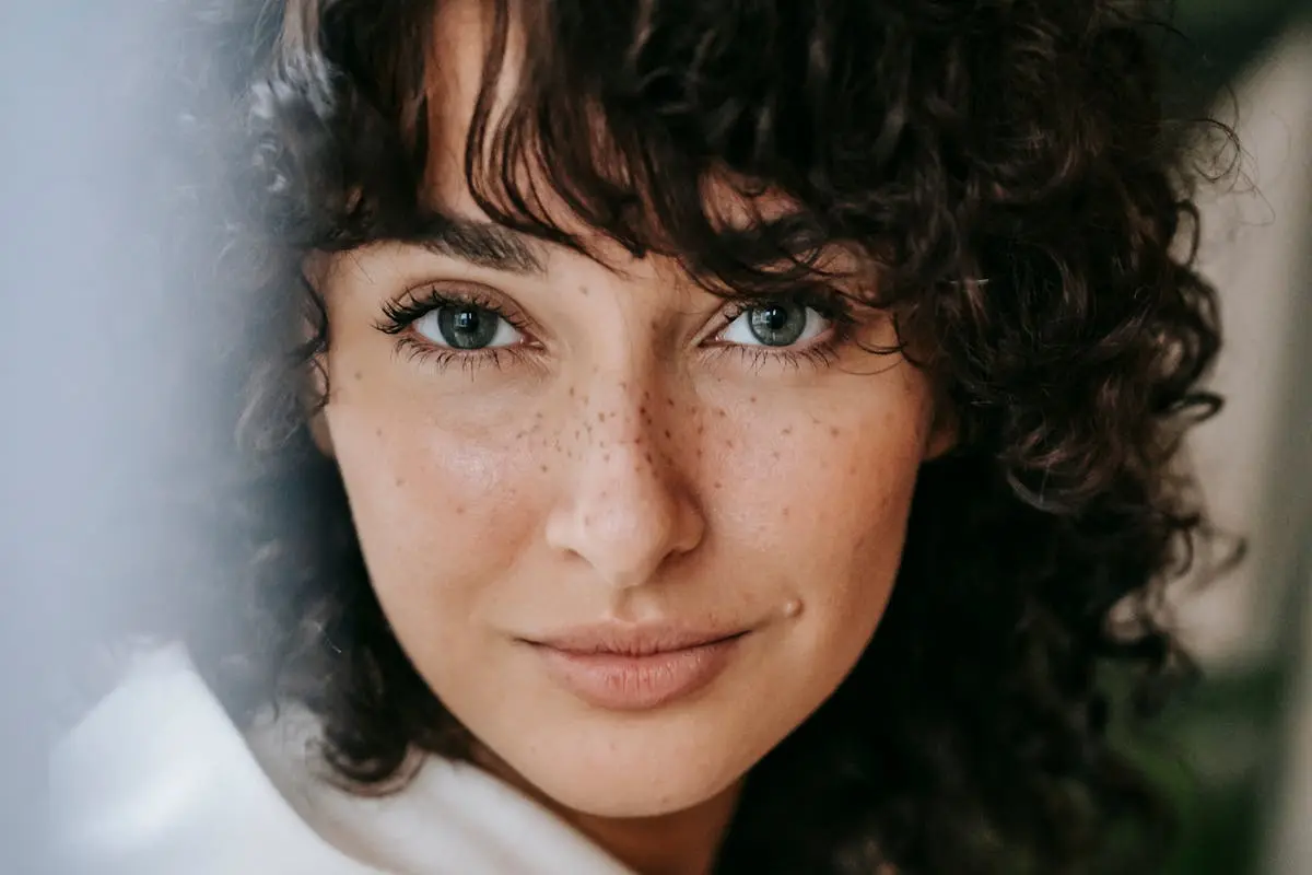 Headshot of attractive female with dark curly hair looking at camera against green leaves on blurred background in light room