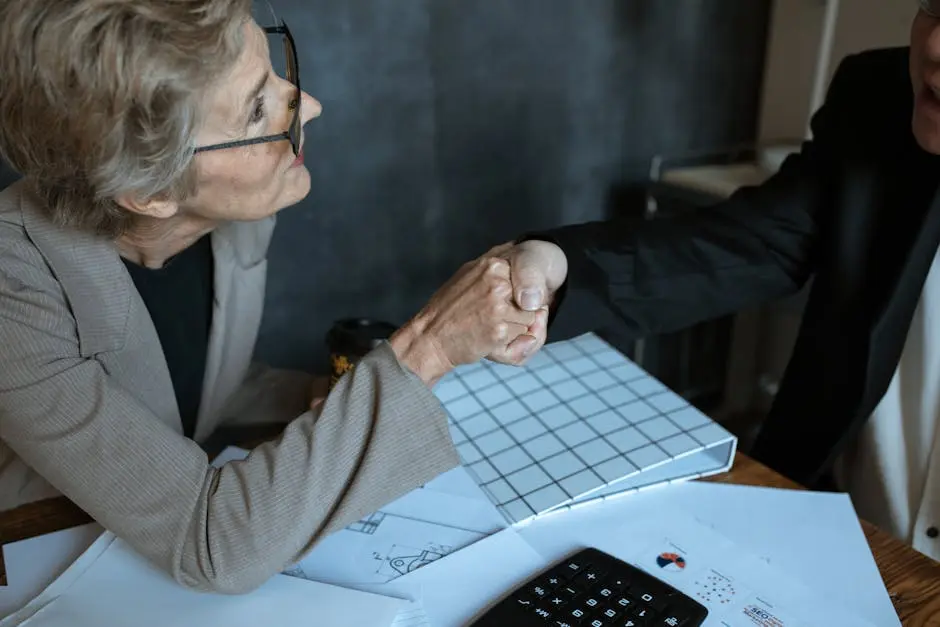 Senior woman in business suit shaking hands across a table in an office environment.