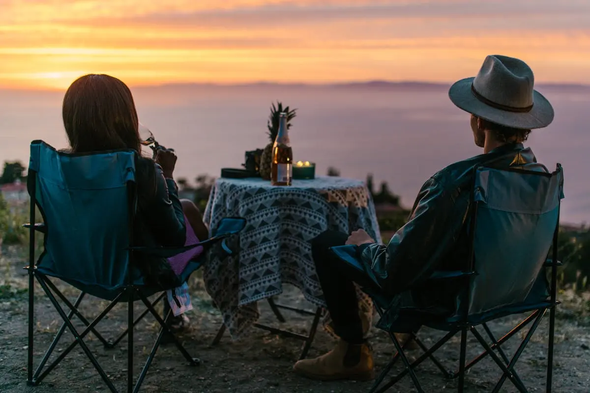 A couple relaxes with drinks outdoors, overlooking a stunning sunset by the sea.