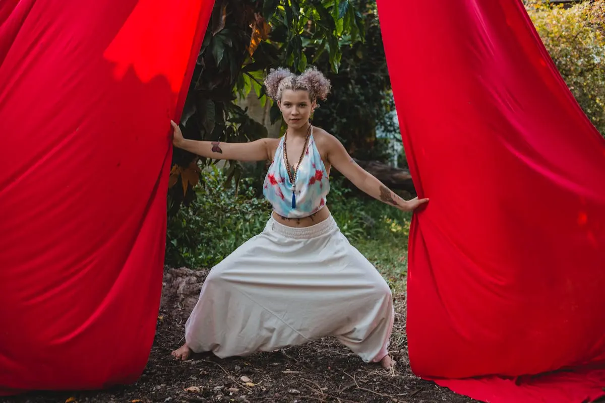 Full body of trendy fit young lady in activewear standing between red aerial silks while practicing anti gravity fitness in park